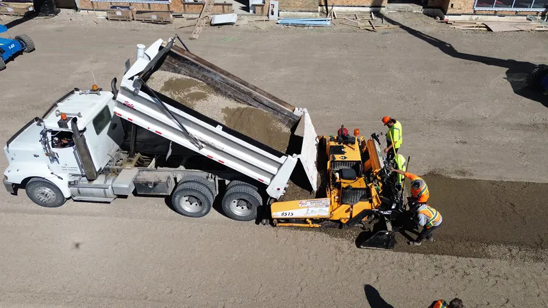 Top-down aerial of a dump truck feeding the paver as the gravel base goes down before asphalt