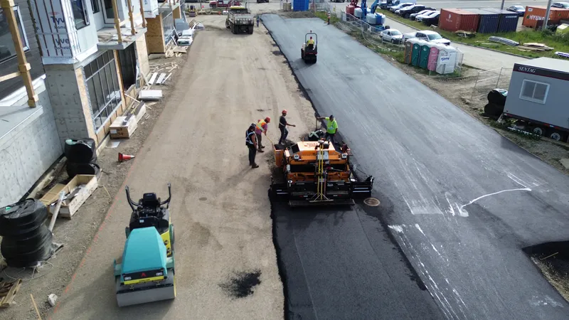 Aerial view from behind the paver with the crew on the deck and a roller compacting the fresh asphalt