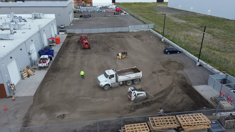Aerial of a fenced compound lot in SE Calgary during base prep: dump truck, skid steer and roller on the graded gravel