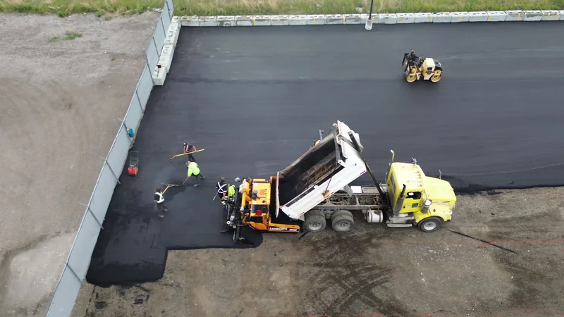Dump truck feeding the paver as the first hot mix asphalt mat goes down along the compound wall